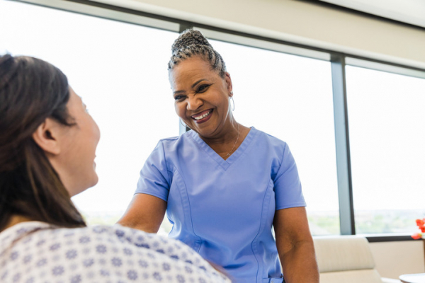 A birth doula in blue top smiles at a pregnant woman seen from the side wearing a hospital gown